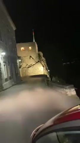 At night, from behind the windshield of a red car, the scene shows a narrow, paved road leading to a stone structure. The structure is lit with two windows glowing in the darkness and a flag on top, presumably illuminated by a spotlight. The structure appears to be built on or into a rocky hill. A tall, light-colored building with multiple windows runs along the left side of the road. Street lights illuminate the road ahead. In the background, a few lights suggest a distant town or city across a body of water. The car's headlights are on, shining up the road toward the structure.