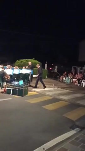 A band is performing outdoors at night for an audience seated in chairs. The band members, dressed in dark uniforms with light blue accents, are standing and playing instruments. A conductor, in a dark suit, stands in front of the band, gesturing with his hands. The audience members, dressed in casual attire, are watching the performance. The scene takes place in front of a building, with trees and bushes in the background. A crosswalk with yellow stripes is visible in the foreground.