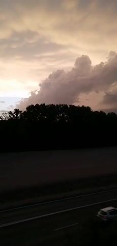 A video shows a dark landscape with a tree line and a road. The sky above is filled with dramatic clouds, some illuminated by sunlight. A few cars can be seen driving on the road. The video appears to be filmed from a moving vehicle.