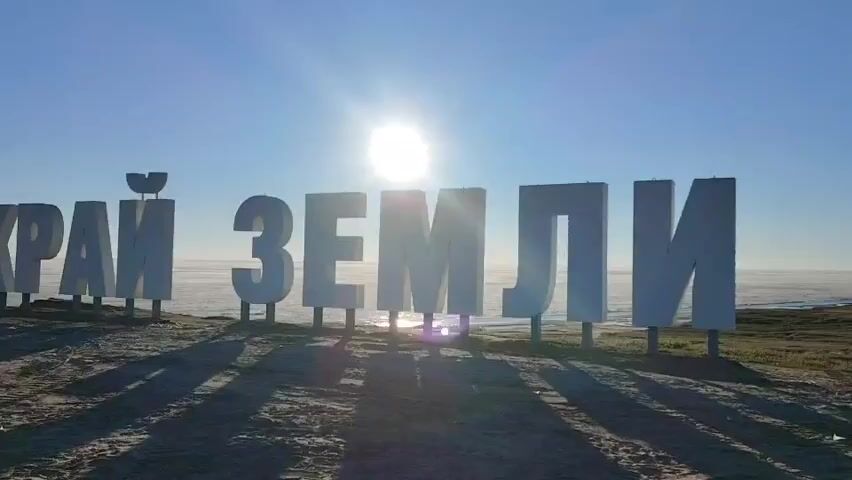 The video shows a large outdoor sign made of white block letters. The sign spells out "КРАЙ ЗЕМЛИ" in Russian, which translates to "End of the Earth." The letters are tall and stand on poles, casting long shadows on the ground in front of them. The sun is low in the sky, shining brightly and creating lens flare in the center of the frame. The background is a hazy, pale blue sky with a faint line of white in the distance, possibly indicating a foggy or misty horizon. The ground is a mix of dirt and what appears to be sparse, dry vegetation. The overall impression is one of a remote, open location, possibly a coastal or elevated viewpoint, bathed in the early morning or late afternoon light. There are no people visible in the video. The camera is static, capturing a wide shot of the sign and its surroundings.