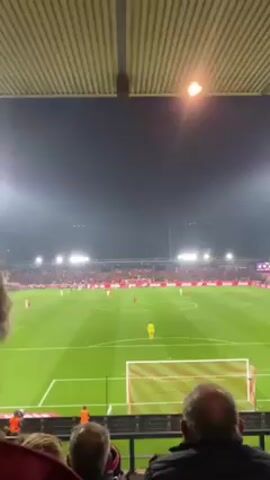 The video shows a soccer game from a spectator's perspective. The field is illuminated by stadium lights, and the sky is dark. Players are visible in the distance, with one standing near the goal. The crowd is densely packed in the stands. The foreground shows the backs of people sitting in front of the camera, with the stadium's ceiling visible above.
