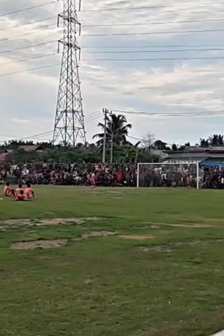 The video shows a soccer match taking place on a grassy field. In the foreground, the field is mostly empty, with some patchy grass. In the background, there is a crowd of spectators gathered behind a soccer goal. A player in an orange uniform is approaching the goal, presumably to take a penalty kick. A goalkeeper in dark clothing is positioned in the goal, ready to defend. The sky is overcast, and a tall power line tower is visible in the distance. Palm trees and buildings are also present in the background. The scene appears to be a casual, local soccer game with a passionate audience.
