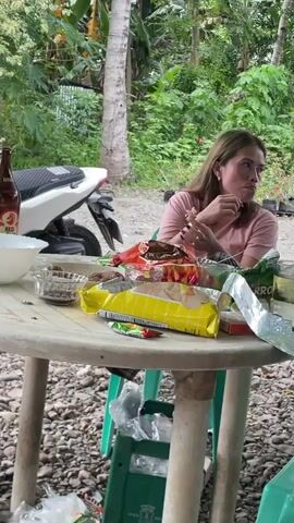 A woman sits at a round, white plastic table outdoors, surrounded by bags of chips and other snacks. She is wearing a pink shirt and has tattoos on her arm. She picks at the snacks while looking around. The table is cluttered, and there is a motorcycle parked nearby. The area is surrounded by lush green vegetation, including trees and various plants. The camera moves up to the canopy of trees above the table.