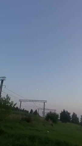 The video shows a time lapse of a sunset over a body of water. In the foreground, there is a grassy hill with some trees and bushes. In the background, there is the sun setting over the water, with a pale pink and orange sky. There are some power lines and industrial structures visible on the horizon in the distance. The video begins with a view of the sky and a crescent moon, then pans across the landscape as the sun slowly descends.