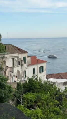 The video shows a scenic view of the Amalfi Coast. Two boats are visible on the blue sea, leaving white wakes behind them. The foreground features houses built on a hillside, characterized by white walls and red tile roofs. Green foliage is abundant, partially obscuring the lower portions of the buildings. A person is visible in the opening of one of the buildings. The sky is clear with some light clouds.