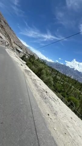 The video shows a person riding a motorcycle on a paved road that winds through a mountainous region. The road is on the left side of the frame, with a steep, rocky mountain on the left and a valley with trees and some buildings on the right. The sky is blue with white clouds. The rider is wearing a helmet with goggles and a mask. The camera is mounted on the motorcycle, giving a first-person perspective of the ride. The video appears to have been filmed on a sunny day.