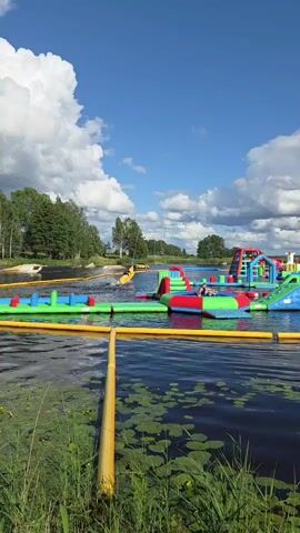 A vibrant inflatable water park is set up on a calm lake under a bright blue sky filled with fluffy white clouds. The park features a variety of colorful structures, including slides, climbing walls, and balance beams, all made of inflatable material. These structures are anchored in the water, creating a playground for water activities. Surrounding the park, the lake is dotted with lily pads, and the shore is lined with green grass and trees, suggesting a natural, outdoor setting. In the distance, a person is visible on a wakeboard, being pulled by a boat, indicating the presence of water sports beyond the inflatable park. The overall scene depicts a recreational area designed for summer fun and outdoor enjoyment.