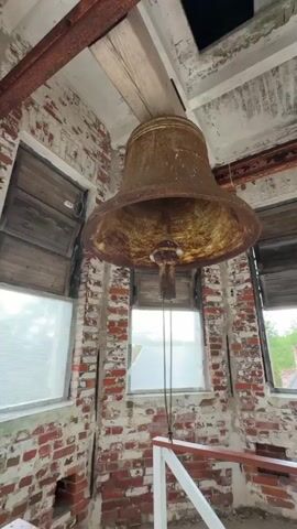 A low-angle medium shot shows a large, rusty bell hanging from the ceiling of a dilapidated structure. The bell is the main focus of the frame. Two ropes hang down from the bell mechanism, one of which is being held by a hand. The walls are made of red brick and are partially covered in white plaster, showing signs of decay. There are wooden shutters and glass windows visible, some of which appear broken. The overall environment suggests abandonment or disrepair.