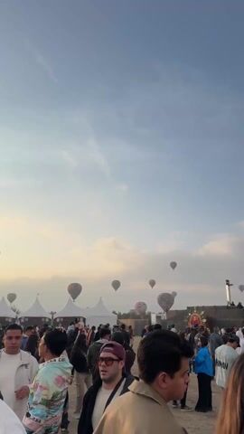 The video shows a crowd of people at a festival or event with a large stage in the background and several hot air balloons in the sky. The camera is positioned behind the crowd, looking towards the stage and the balloons.  The stage has a large screen displaying text (ZONA VIP) and lights. There are people on the stage. Tall speaker towers flank the stage. The hot air balloons are colorful, with various patterns. In the distance, a pyramid-like structure is visible on the horizon. The sky is blue with scattered clouds. Some people in the crowd are looking at the balloons and stage, while others are taking pictures with their phones.