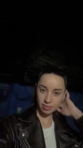 A young woman stands in front of the camera, wearing a brown leather jacket over a white t-shirt. She has her hair pulled back and sunglasses are perched on top of her head. She is looking at the camera, smiling slightly and puckering her lips. The background is dark, with a blue wall partially visible. Branches are briefly seen at the top of the frame.