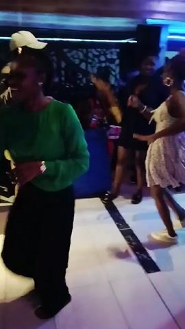 The video shows people dancing in a dimly lit nightclub. The club features a marble-like patterned floor, dark walls, and a blue cushioned booth along one wall. There are multiple people, some dancing and others sitting, and they are dressed in casual clothing. The lighting includes green and red accents, and the camera moves around the scene, capturing the energy of the event. Music is playing in the background.