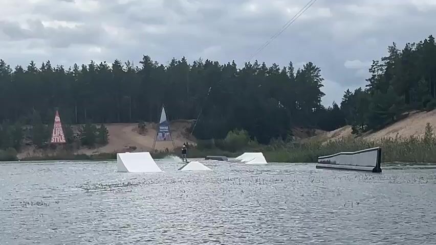 A wakeboarder is seen being pulled by a cable across a body of water. They are surrounded by various obstacles, including ramps and a ledge, set up for wakeboarding. The background features a sandy embankment and a dense forest of pine trees under a cloudy sky. Two triangular banners with text are visible on the embankment to the left of the wakeboarder. The water is choppy, indicating movement.
