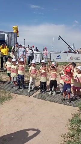 A group of young children, all wearing yellow shirts with bibs and white hats, are standing in a line outdoors on what appears to be a track or field. They are performing a synchronized stretching exercise, bending their bodies to the side with their arms raised. In the background, there are adults, tents, a large inflatable character, and a camera crane, suggesting an event or competition. The sky is blue with some clouds.