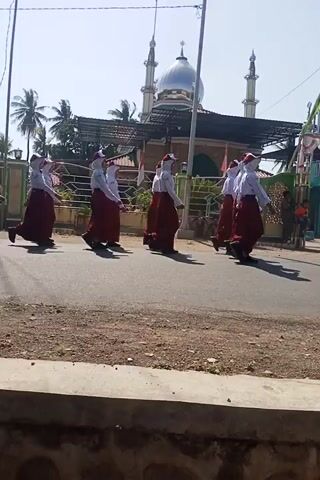 The video shows a group of children in uniform, likely performing some kind of coordinated movement or dance in a street. They are dressed in white tops and maroon skirts or dresses, with some wearing headscarves. The children are arranged in a line, facing away from the camera, and appear to be extending their arms outwards. In the background, there is a building that looks like a mosque with a dome and minarets, and a sign that reads "MASJID FATHOR RAHMAN". The environment appears to be an outdoor street with a road and a gutter. The lighting suggests it is daytime with a clear sky. The camera is positioned at an angle, making the scene appear vertical.