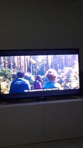 The video shows a television displaying a scene of three people in a forest. The person in the foreground is a young black woman with braids, wearing a purple jacket. Two other individuals, a young red-haired man and another, are slightly behind her, all walking in the same direction. The forest is visible in the background with tall trees and sunlight filtering through the leaves. Subtitles in Bulgarian are visible at the bottom of the screen. The TV is placed on a white cabinet. The room is dimly lit.