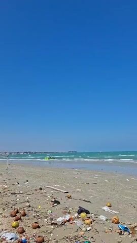 The video begins with a view of a clear blue sky and the ocean. The foreground shows a sandy beach littered with debris, including coconuts, plastic bags, and other unidentifiable trash. The ocean is calm with small waves. As the video progresses, two fishing boats are revealed grounded on the beach. A young person, wearing a light-colored uniform with shorts and a short-sleeved shirt, is seen standing on the beach in front of a larger, colorful fishing boat. The person is holding a kite string and looking up at the sky as a white kite is visible flying. The larger boat is partially on land, supported by a wooden structure. The scene suggests a sunny day at a somewhat neglected beach with active fishing vessels.