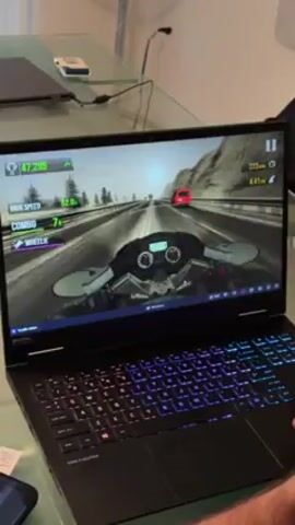 The video shows a person playing a motorcycle racing game on a laptop. The laptop has a black chassis and a multi-colored backlit keyboard. The game is displayed on the laptop screen, showing a first-person perspective of a motorcycle racing on a highway with mountain scenery. The game interface shows the player's speed, combo level, and score. The person is using the trackpad on the laptop to control the motorcycle in the game. The laptop is sitting on a glass table, and there are other objects visible in the background, including a second laptop, a mouse, and a smartphone. The background is a home interior.