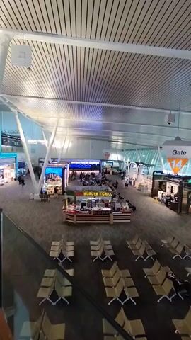 The video shows an aerial view of an airport terminal. Rows of empty, tan-colored seats are arranged on the dark grey floor. In the background, a "Gate 14" sign is visible, hanging from the ceiling. There are several concessions and shops on either side of the terminal, including a "Burger & Grill" stand in the center. A few people are scattered throughout the terminal, walking and standing. The ceiling is made of white, slatted panels, and large, white support beams are visible throughout the space. Large windows line the far wall, offering a view of what appears to be an airfield. The lighting is bright and artificial. The video pans slightly to the right.
