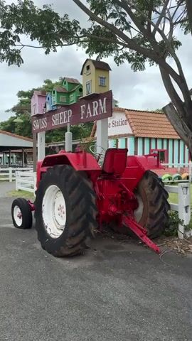 The video pans from left to right, showcasing a farm scene. In the foreground, a bright red tractor with large tires is parked on an asphalt path. Behind the tractor, a wooden sign reads "SWISS SHEEP FARM" with smaller text below indicating "Horse Station". The sign is supported by white posts and is adorned with small, colorful, house-shaped decorations. Further in the background, a white building with red shutters and two blue windows is visible. To the left of the building, a white fence encloses a grassy area with large, white sheep sculptures. In the distance, a white windmill stands tall, and a variety of trees and structures suggest the layout of the farm. The sky is overcast with clouds.