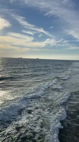 A vertical video shows a vast ocean with choppy waves under a partly cloudy sky. The sun, partially obscured by clouds, shines brightly on the water's surface, creating a shimmering reflection. In the distance, several large ships are visible on the horizon. The camera perspective is from a pier or elevated platform, with a portion of a light blue railing visible in the lower left and bottom center of the frame.