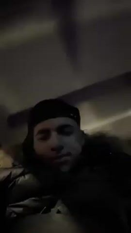 A low-angle video features a young man wearing a black beanie and a dark, fur-trimmed hooded jacket. He sits back against something indistinct, looking straight at the camera with a slight smile. The lighting is dim, causing shadows to play across his face and the environment. As the music starts, he raises a hand and obscures the lens momentarily, revealing a glimpse of someone seated next to him before returning the focus to himself.