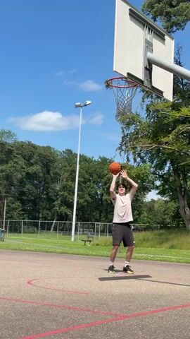 A teenage boy stands on a basketball court, preparing to shoot a basketball. He is wearing a light-colored t-shirt, dark shorts, and athletic shoes. He holds the basketball with both hands in front of his chest, his eyes focused on the hoop. The basketball court is outdoors, with a red boundary line visible on the ground. In the background, there are trees and a clear blue sky with some clouds. A basketball hoop with a net is positioned above and to the right of the boy. The boy shoots the ball, and it flies toward the hoop. The ball goes through the hoop. He then catches the ball after it falls through the net. He then shoots the ball again, and this time it misses the hoop. He catches the ball and prepares to shoot again.