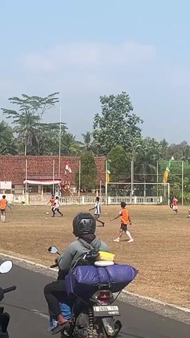 The video shows an outdoor soccer game being played on a dry, grassy field. Several young men in athletic attire are visible on the field, some actively playing soccer and others observing. One person is wearing an orange jersey with the number 9, and another is in a green jersey with the number 9. In the background, there is a building with a red roof and flags, possibly a school or community center. Trees and palm trees surround the area, suggesting a warm climate. A motorcycle is parked in the foreground on the left side of the frame, and a person is sitting on it, partially obscuring the view of the soccer game. The sky is clear and blue, indicating a sunny day. The overall scene depicts a casual soccer match taking place in a rural or suburban setting.