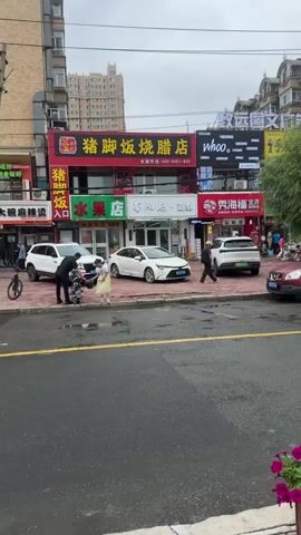 The video shows a street scene with several cars parked and driving. Buildings line the street, with signs visible in Chinese characters. There are a few people walking on the sidewalk. The weather appears overcast. In the foreground, there are planters filled with pink and purple flowers. A white sedan is parked on the left side of the street, with a person in a black jacket standing next to it. A white SUV is parked further down the street, and a person in dark clothing is standing near it. A dark-colored SUV is driving past in the foreground. A red SUV is parked on the right side of the street. A white van is parked behind the red SUV. A white box truck is driving down the street. A gold-colored taxi is driving in the distance. Several tents or canopies are set up along the sidewalk.