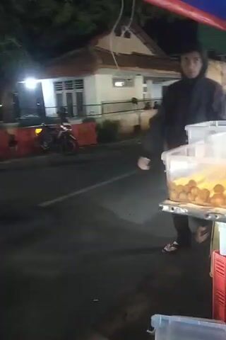 The video shows an outdoor scene at night, likely a street food stall. In the foreground, there are transparent plastic containers filled with food items, some appearing to be on sticks. A person wearing a dark hooded jacket and dark pants, along with sandals, walks past the food containers. In the background, other people can be seen, and there are signs and lights, indicating a busy commercial area. The overall impression is that of a food vendor at a night market or similar setting.