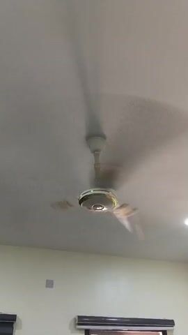 The video shows a room's interior, focusing on the ceiling. Initially, a white and gold ceiling fan is rotating. The camera then pans to show a recessed LED light in the ceiling. The walls are a light beige color. A portion of a window with a dark shade is visible at the bottom of the frame.