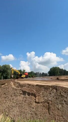 The video shows a construction site under a bright blue sky with fluffy white clouds. A large yellow excavator is parked on a mound of dirt on the left side of the frame. Next to the excavator is a large orange tank. In the middle of the frame, two people are visible. One person appears to be walking, while the other is operating a hose that is spraying water onto the ground. The ground is covered in dirt and what appears to be a white sheeting material. Trees and vegetation are visible in the background. The overall impression is of ongoing construction work in an outdoor environment.