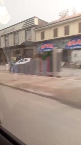 The camera travels down a city street, capturing storefronts and passing vehicles. Buildings are primarily one or two stories tall, with signs in a language other than English. Cars, including a white hatchback, a silver sedan, and a blue van, are seen parked or moving along the street. The environment appears urban, with a cloudy or hazy sky overhead. The video is taken from a moving vehicle, giving a slightly shaky perspective of the scene.