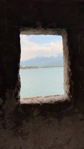The video shows a view of Lake Geneva in Switzerland, seen from a stone window frame. The lake is calm and blue, with a few small boats visible. In the background, the Alps rise majestically, their peaks covered in snow. The sky is blue with scattered white clouds. As the camera pans slowly outward, it reveals the full vista of the lake and mountains.