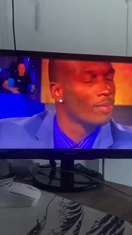 The video shows a computer monitor displaying a television program. The main subject on the monitor is a Black man wearing a blue suit and a blue striped shirt. He is looking directly at the camera and appears to be speaking. In the upper left corner of the monitor, a smaller video feed shows a White man with short hair, wearing a black shirt, sitting in a dimly lit room with blue and purple lighting. He is gesturing with his left hand. The television screen has the brand name "PHILIPS" visible at the bottom. The monitor is placed on a desk or table, and in the foreground, a white computer mouse and a patterned mousepad are visible. The background behind the monitor is a plain wall.