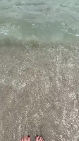 A video shows the gentle lapping of clear, shallow ocean waves onto a sandy shore. The water is transparent, revealing the rippled sand beneath. In the distance, a sailboat is visible on the horizon under a hazy sky. The camera is positioned close to the water's edge, capturing the movement of the waves and the texture of the sand.