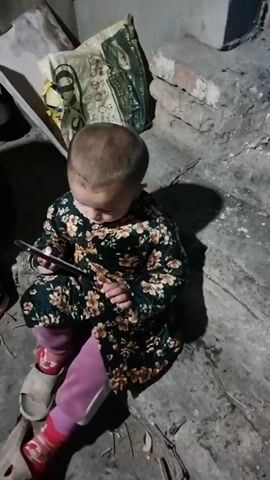 A young child is sitting on a concrete floor, holding a white smartphone. The child is wearing a floral print robe, pink leggings with red and white striped socks, and tan sandals. The background includes a bag with green and yellow designs, and some broken concrete. The lighting is dim, casting a shadow of the child. The child is looking upwards with a slightly concerned expression.