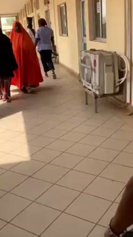 The video shows an indoor hallway with beige tiled floors. People are walking in the hallway. Initially, two women are seen walking, one in a black hijab and one in a large, orange wrap. As the camera moves, other people are visible, including individuals in medical scrubs and casual clothing. An air conditioning unit is visible on the side of the hallway. Towards the end of the video, a man in jeans, a grey shirt, and a white baseball cap is looking out over a balcony. Trees are visible outside.