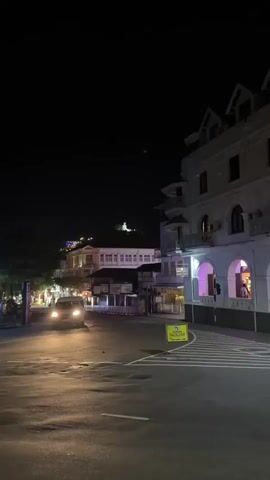 The video depicts a night scene in what appears to be a city street. The camera tilts upwards to focus on a large, illuminated statue of Buddha situated on a hilltop in the distance. In the foreground are buildings, a street with white striped crosswalk, a yellow sign on the side of the street, and a car with its headlights on, driving on the road. The overall lighting is dim, with the main source of light coming from streetlights and the illuminated statue.