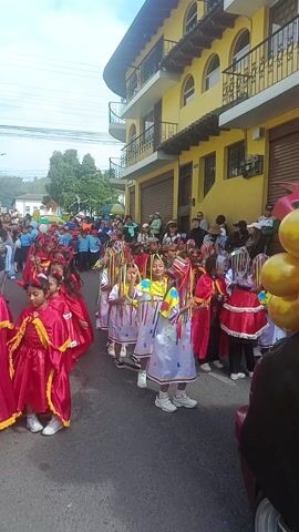 The video captures a lively street parade or procession featuring numerous children. The scene is set outdoors on a paved street, lined by buildings. A prominent two-story building on the right side of the street features bright yellow walls, dark-colored window frames, and balconies. In the background, more buildings and some green foliage can be seen, with power lines crisscrossing overhead.

The children are the main focus, dressed in colorful costumes. Many of the children, particularly on the left and right sides of the frame, are wearing bright red capes or dresses, some adorned with gold trim. In the center of the procession, several children are dressed in white tunics or dresses that feature colorful, abstract shapes (yellow, blue, red) on their fronts. These children also wear unique headpieces from which long, multi-colored ribbons (yellow, green, blue, red) stream down, framing their faces and swaying as they move.

The children are walking forward in an organized manner, some appearing to dance or sway to the music. Their movements make the colorful ribbons on their headpieces flutter. Adults and other spectators line the street, observing the procession. A partial view of a yellow balloon arrangement is visible on the far right. The sky is bright, indicating it is daytime. On the left side of the street, a blue sign with white text reads "Alianza del Valle".

The atmosphere appears festive and joyful, characteristic of a community or cultural celebration. Traditional-sounding music with percussion and wind instruments plays in the background, adding to the celebratory mood.