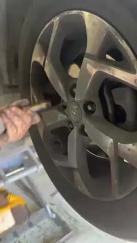 The video shows a close-up of a car wheel, which is dirty, with a grey rim, secured on a lift in a garage. The wheel has a complex spoke design, and the central hub is visible. Behind the wheel, the brake rotor and caliper are noticeable. The lift mechanism is partially visible at the bottom left, and the floor appears to be concrete. The lighting is somewhat diffused.