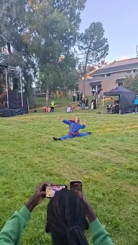 A person in a blue jumpsuit with red fringe and sunglasses is doing a split on a grassy lawn. They have their arms raised. Behind them, several people are standing and watching, some dressed in period clothing. There's a stage setup to the left and a building in the background. The scene appears to be outdoors during the day. The bottom of the video shows two hands holding a smartphone, filming the performer.