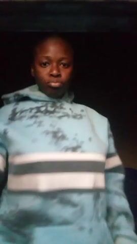 A young woman with short hair stands in a dark room, wearing a light blue hooded sweatshirt with white and gray stripes across the chest. The sweatshirt appears to have dark spots. She makes a gesture resembling a salute with her right hand, then moves her arms. She turns her head to the side and looks back at the camera with a serious expression. She is also wearing pink bottoms.