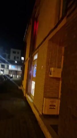 The video shows a building at night, decorated with Christmas lights that change color. The building is made of brick and has a window. There is a sidewalk in front of the building, and across the street there are other buildings and cars parked. The camera moves slightly while recording.