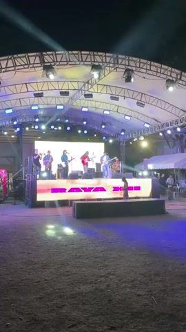 A band is performing on a brightly lit stage outdoors at night. The band consists of at least five members, most of whom are playing guitars. A large LED screen is behind the band, displaying the word "RAVAGE" in pink letters. Several spotlights are positioned above the stage, illuminating the performers and the surrounding area with colorful lights. The ground in front of the stage is covered in dry grass and dirt. A few people are visible in the background, including one person standing near the stage and another person walking further away. There are tents and structures visible in the distance. The overall atmosphere appears to be a lively outdoor concert or event.