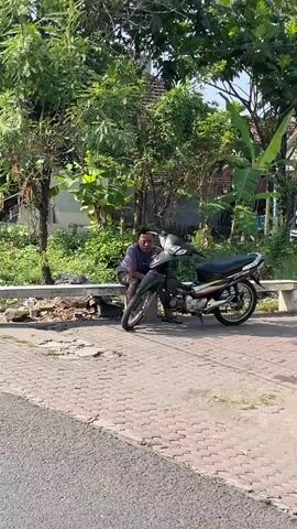 A man sits on a concrete ledge next to a motorcycle parked on a paved area. Trees and foliage surround the scene, with a house visible in the background. The man is wearing a blue t-shirt and dark shorts. The motorcycle is black with yellow and red accents. The weather appears to be sunny.