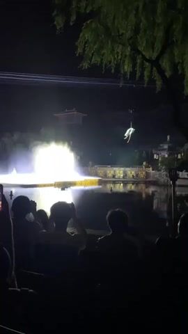 The video shows an outdoor nighttime performance. A brightly lit stage is on the water.  An aerialist is suspended in the air.  People are silhouetted in the foreground, some are holding up phones to record the show. The background shows buildings and trees. The lighting is dramatic, with bright spotlights on the stage and the aerialist.