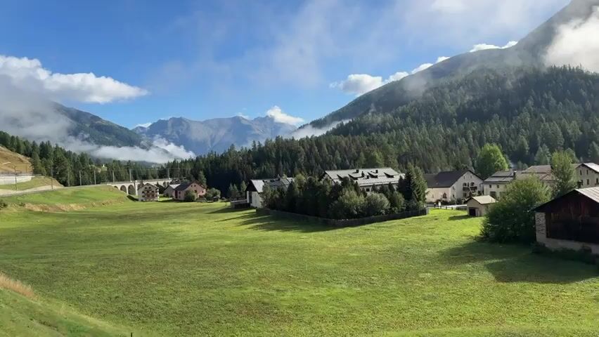 A panoramic view captures a serene valley landscape under a partly cloudy blue sky. In the foreground, a lush green meadow stretches across the frame, bordered on the left by a fence and a road winding up a hillside. A red train passes by on the left side of the frame. In the middle ground, several quaint houses with pitched roofs are nestled amongst trees, forming a small village. An arched stone bridge is visible in the distance on the left. The background is dominated by towering mountains, with dense green pine forests covering their lower slopes and clouds clinging to the peaks. The overall scene depicts a picturesque rural setting in a mountainous region.