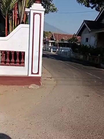 The video shows a street scene in what appears to be a rural or semi-rural area. On the right side of the street are houses and vegetation. A blue van is parked in an open garage area under a building with a banner that reads "AN NASTAS KUE KERING," suggesting it is a shop selling dry cakes. Further down the street, there is a flagpole. On the left side of the street, there are more houses and trees, with a distant mountain visible in the background. The road is paved, and there are no visible people in the frame. The lighting suggests it is daytime.