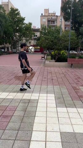 A man juggles a soccer ball in an urban environment. He stands on a tiled plaza made of grey, red, and white tiles. The man is wearing a black t-shirt with a cartoon character print, black shorts, and black shoes with green socks. He juggles the soccer ball with his feet and knees, maintaining his balance. In the background, there are trees, park benches, buildings, and a red food truck. The weather appears overcast. The camera follows the movement of the ball, sometimes tilting downwards to show the tiled surface.