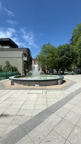 The video shows a round, tiered fountain in operation during the day. Water is being forcefully projected upward from the center of the fountain, creating a visible spray. Additional water spouts are situated along the concentric rings within the fountain, arcing water downward. The fountain is constructed with light-colored stone or concrete. The surrounding environment includes buildings with modern architecture, trees with green foliage, and a paved walkway of light-colored tiles or stones. The sky is mostly blue with some visible clouds. The overall scene conveys a sense of urban tranquility.